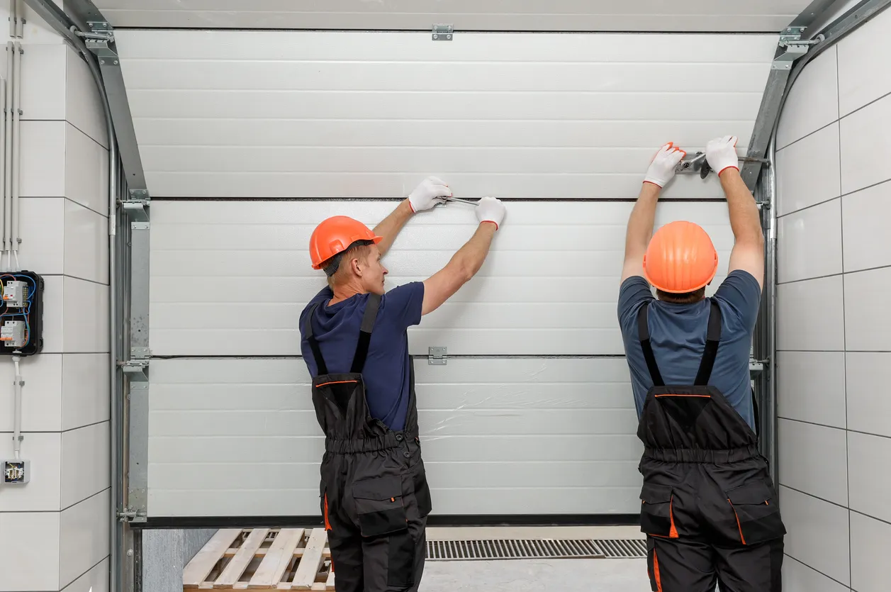 Two workers wearing orange hard hats and dark overalls are standing on a platform, using tools to install a white sectional garage door in a tiled workshop or garage