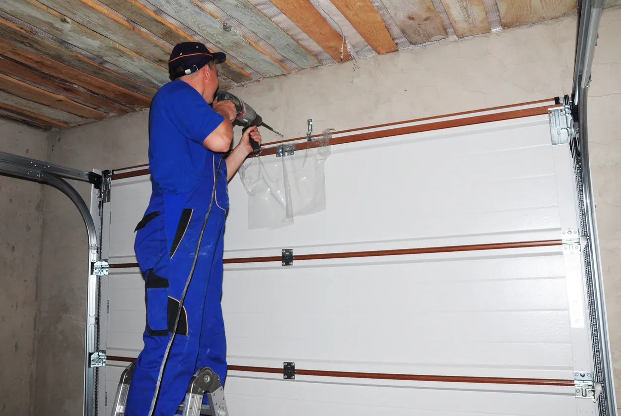 A man in a blue jumpsuit and baseball cap uses a power drill to install hardware on a white sectional garage door while standing on a ladder.