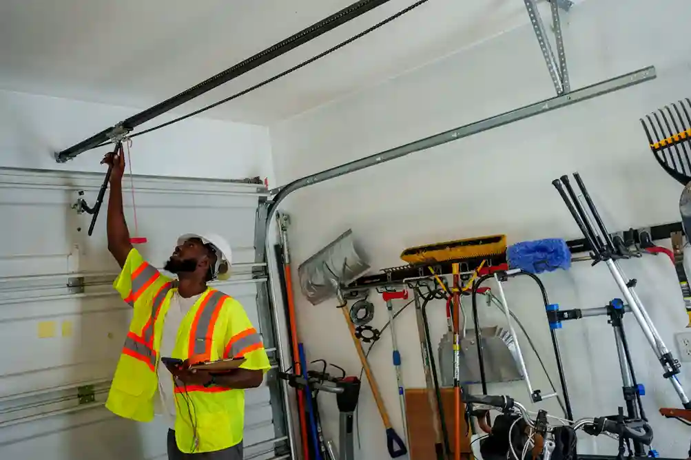 An African American man wearing a white hard hat, safety vest, and holding a tablet, is performing maintenance on the chain-drive system of a garage door.