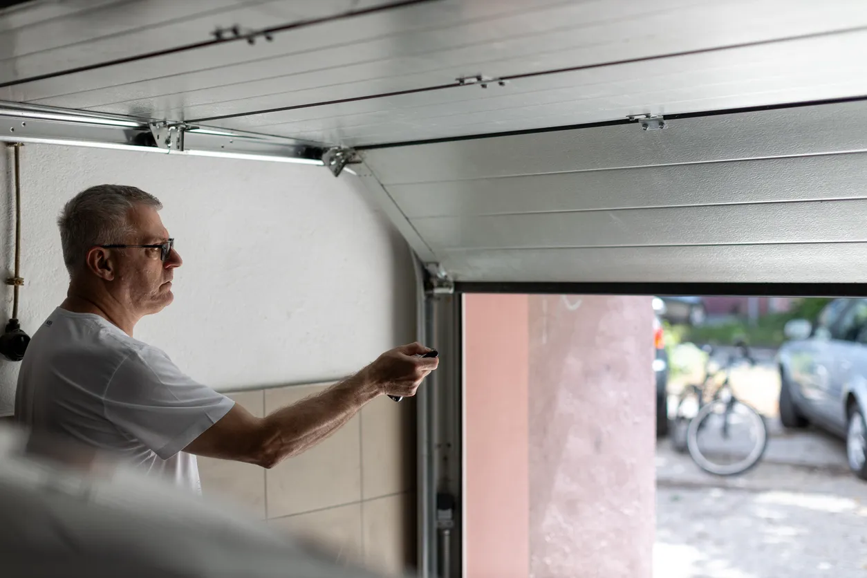 An older man with glasses and a white t-shirt is using a garage door remote to open a white sectional door from inside his garage.
