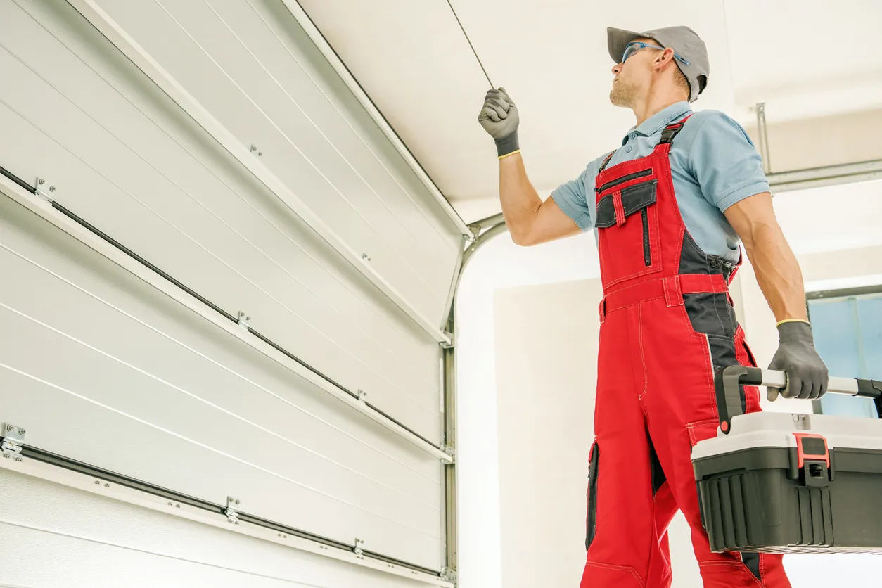 A professional technician in red overalls and a grey cap is performing maintenance on a garage door, holding a tool and a toolbox.