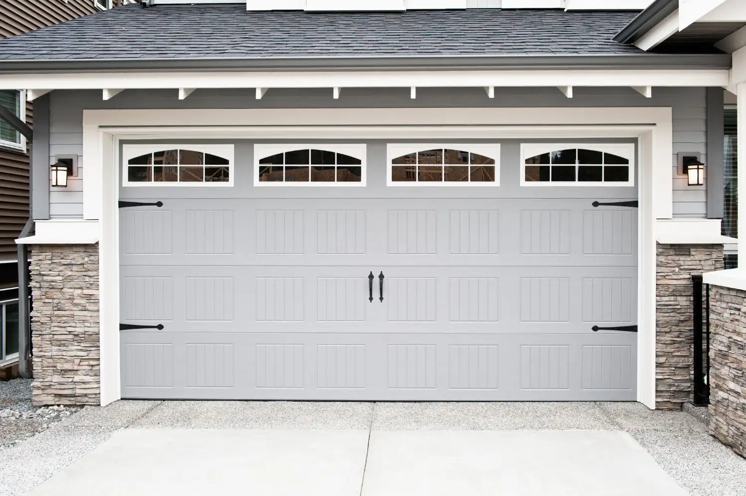 A modern light gray house features a matching gray garage door with four arched windows and black decorative hardware, including handles and hinges.