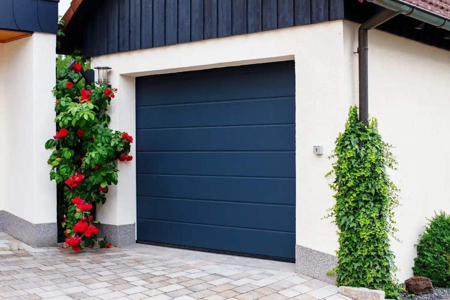 A single dark blue horizontally paneled garage door is set into a light cream-colored house with dark wood siding, with red roses climbing up a white pillar on the left.