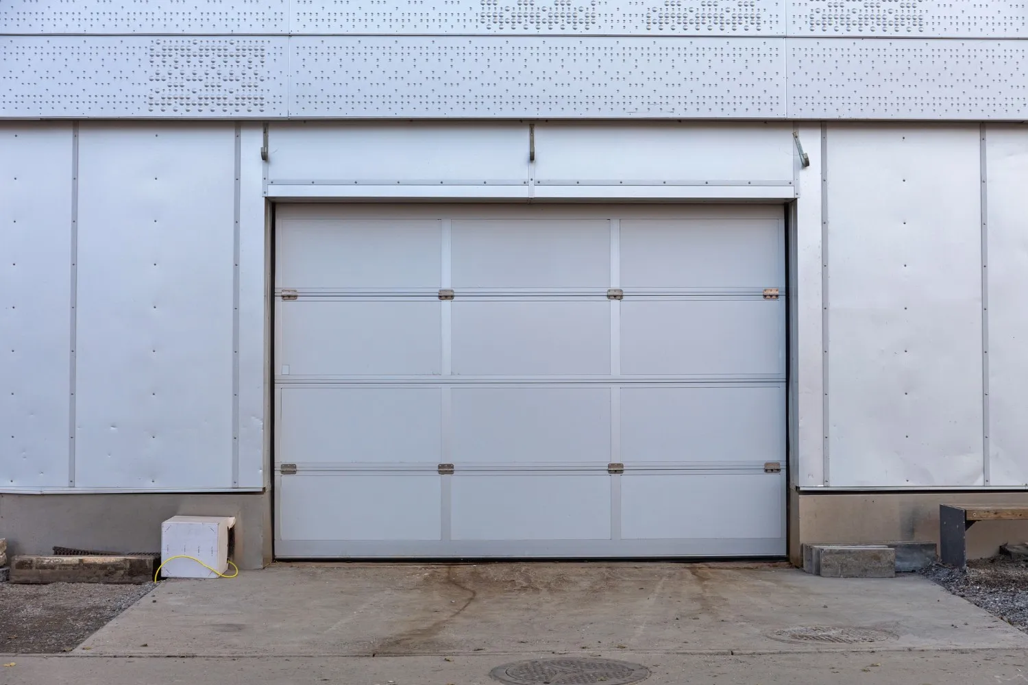 A wide, white garage door with horizontal panels and small gray latches is centered on a light gray industrial building with a concrete driveway leading up to it.