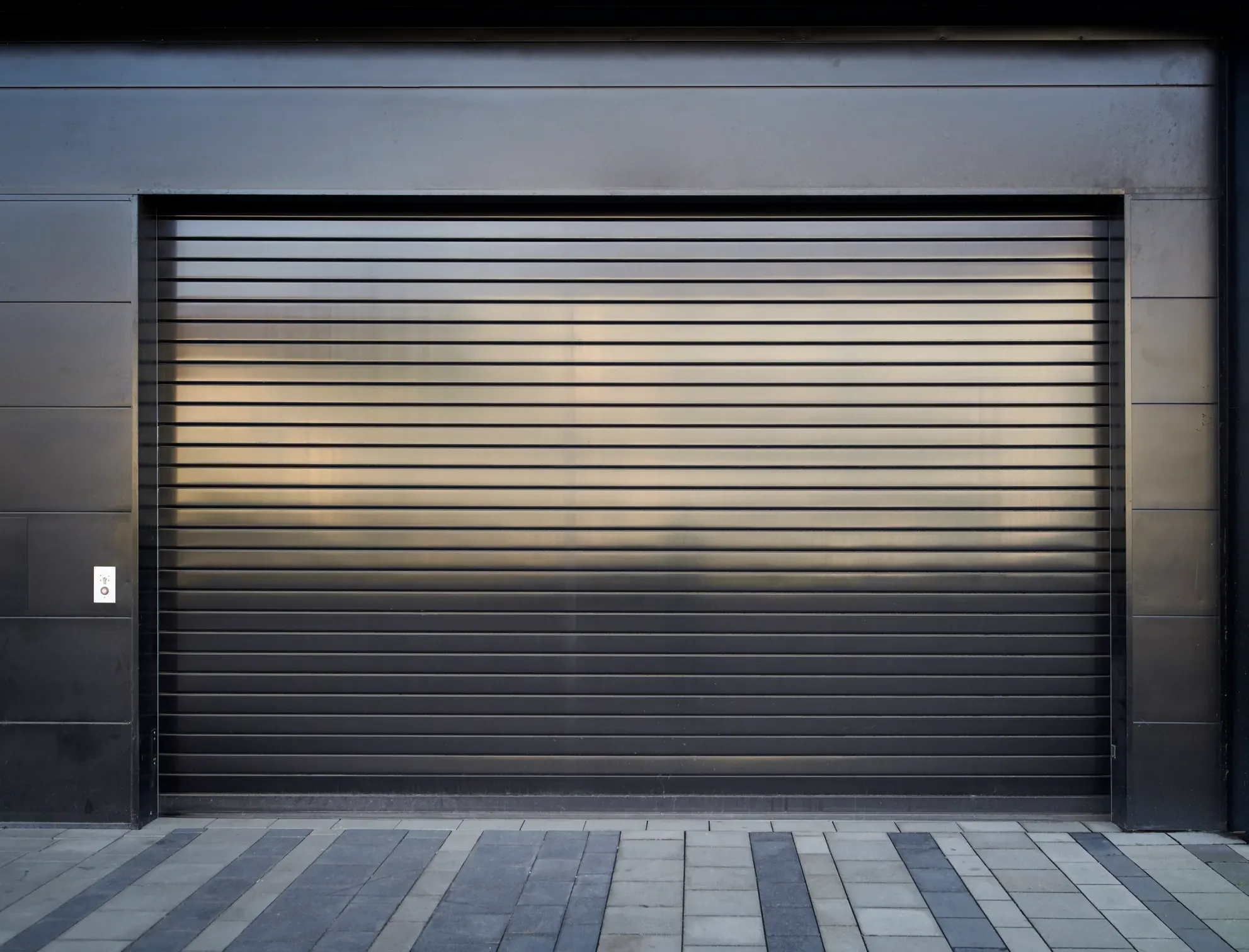 A black, horizontal slat-style rolling garage door on a modern building, with a small doorbell on the left.