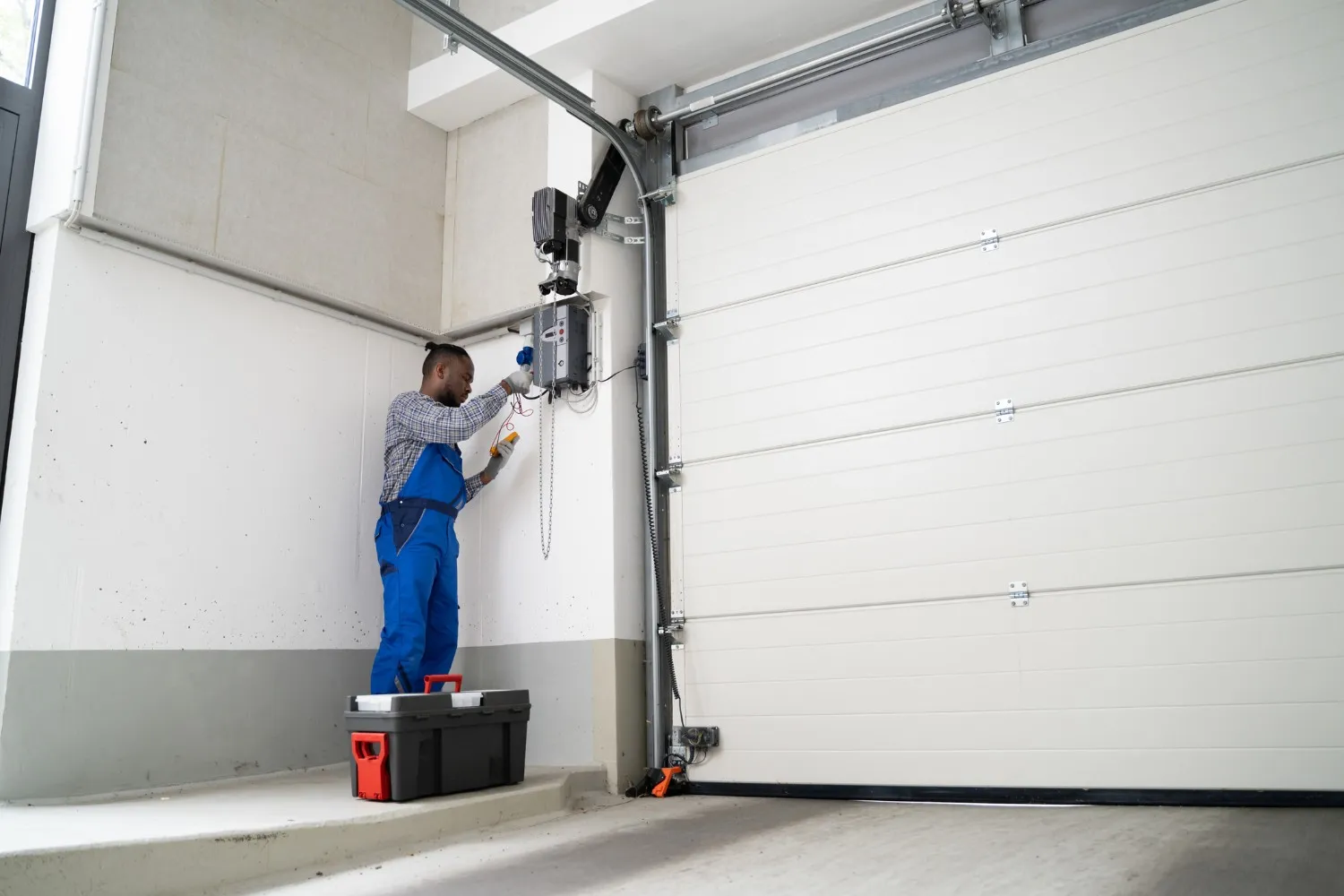 A technician in blue overalls is standing on a toolbox, working on the motor and wiring of a white garage door opener mounted above a large, white paneled garage door.