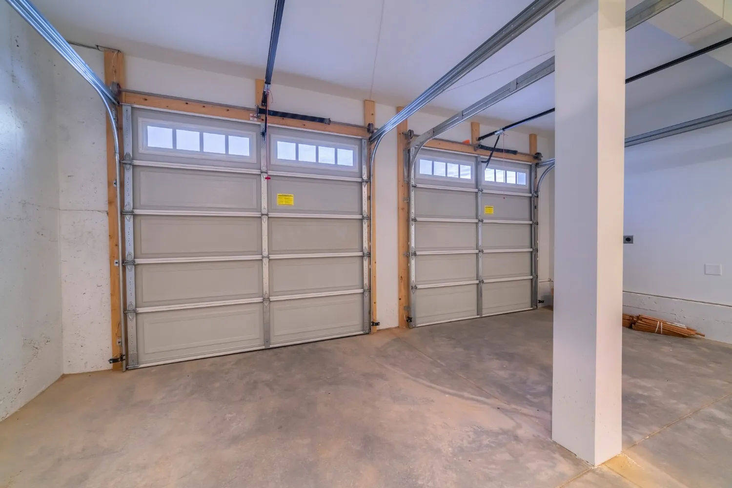 An empty two-car garage features two light gray paneled garage doors with windows at the top, a concrete floor, and white walls with exposed wooden beams and metal tracks.