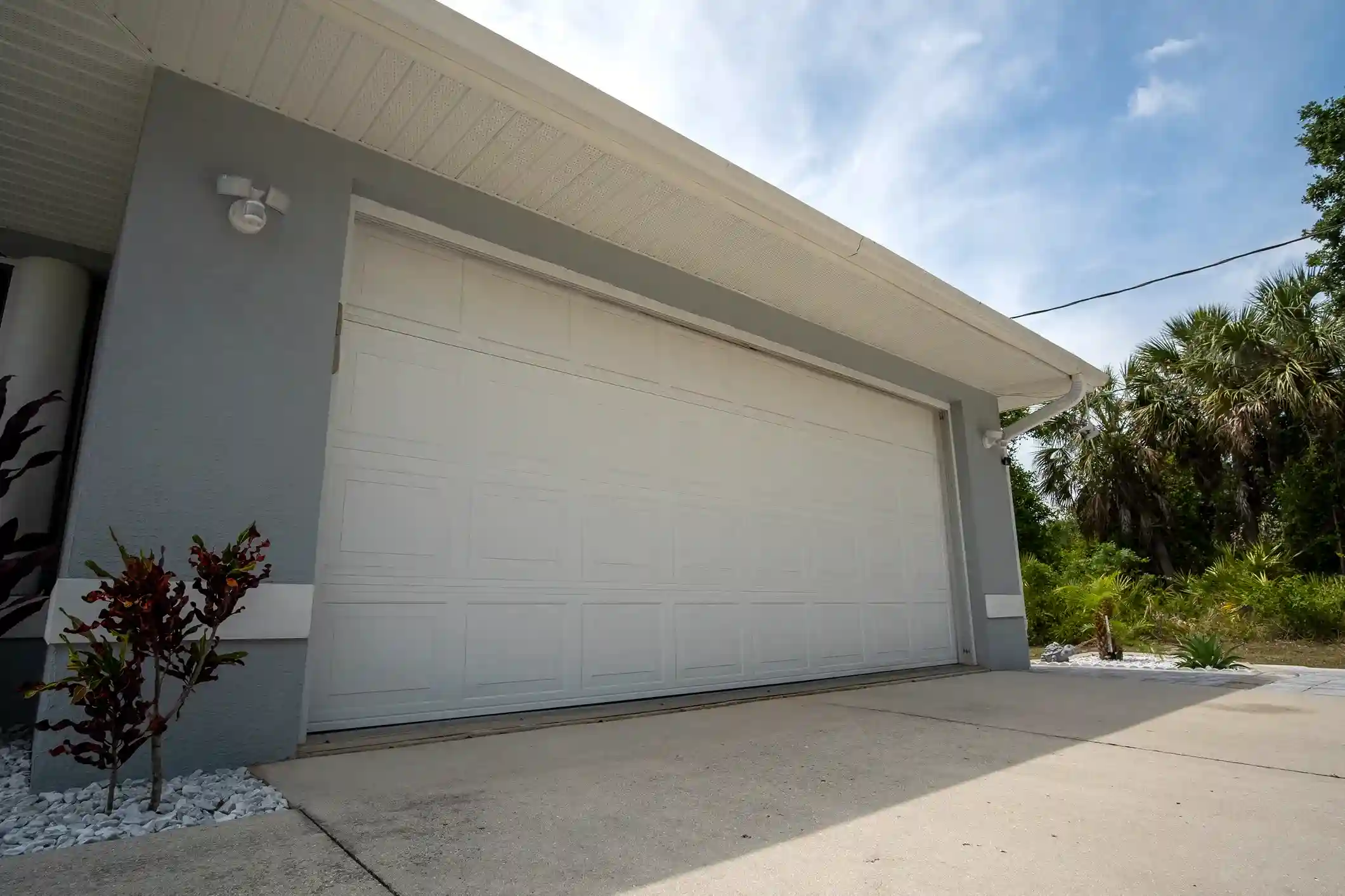 A large, simple white paneled garage door on a gray stucco house, with palm trees and foliage visible in the background.