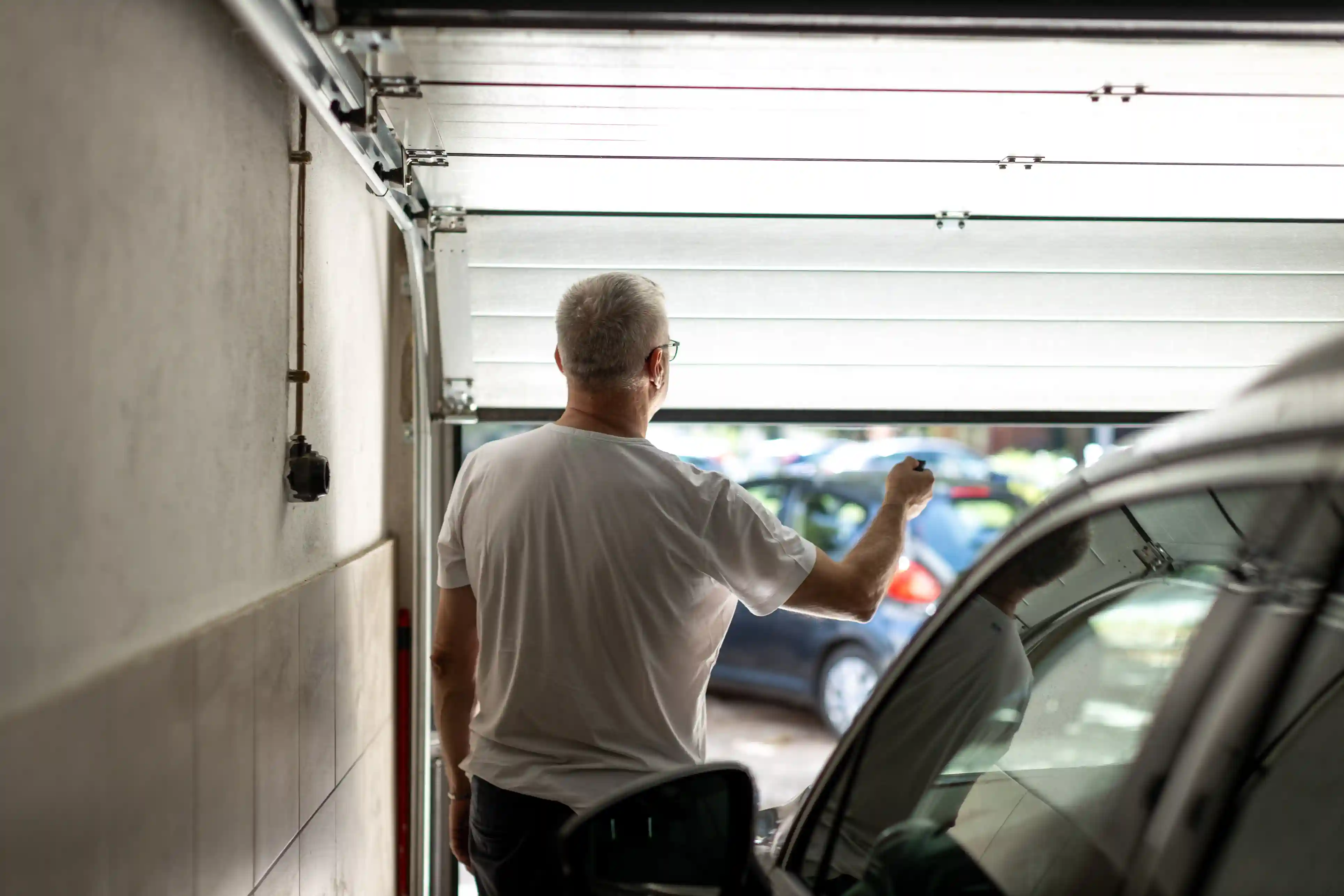 A man in a white t-shirt from behind, reaching up to open a garage door from the inside, with cars visible outside.