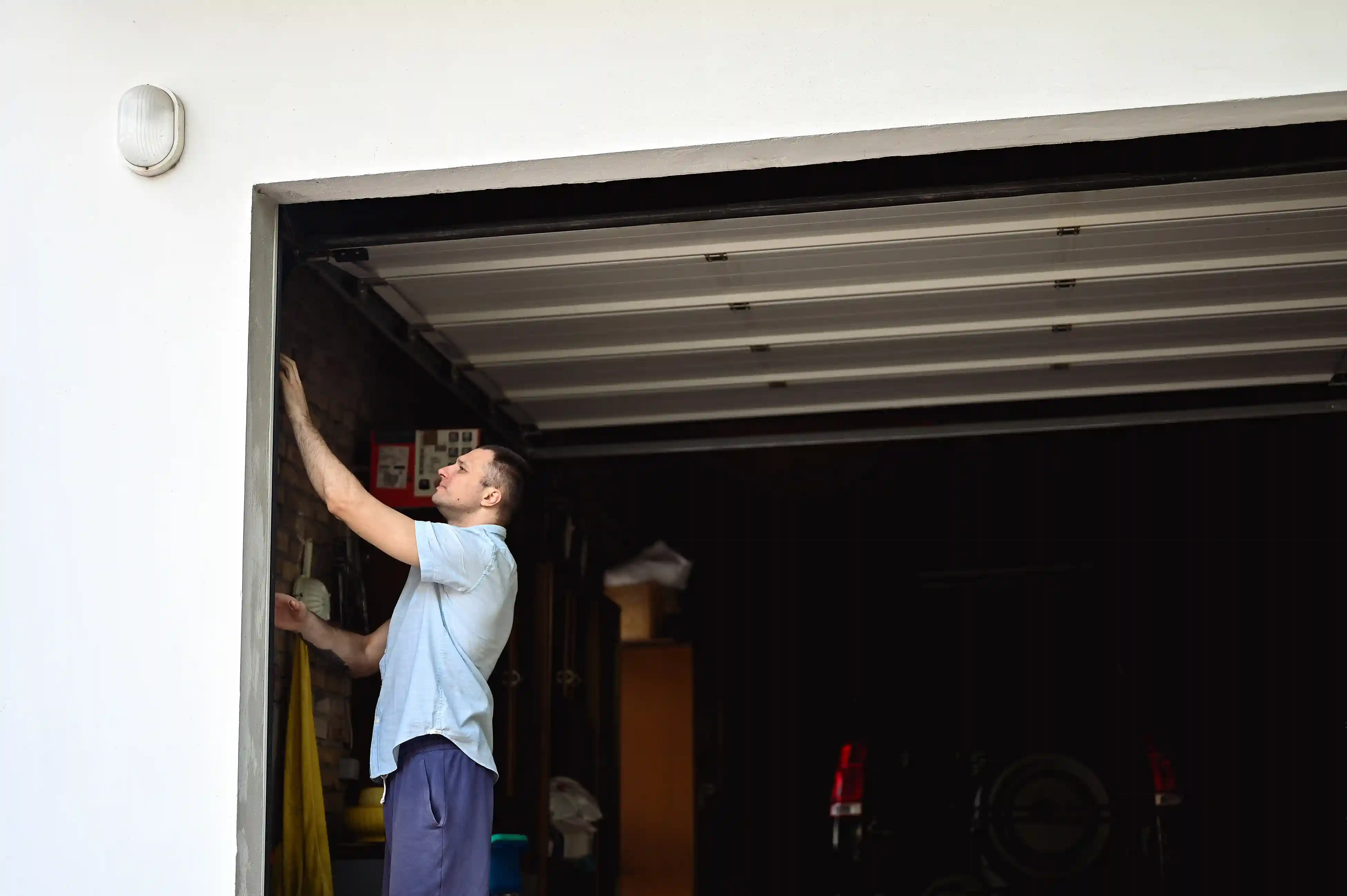A man in a light blue shirt adjusting the side mechanism of an open garage door, with the dark garage interior visible.