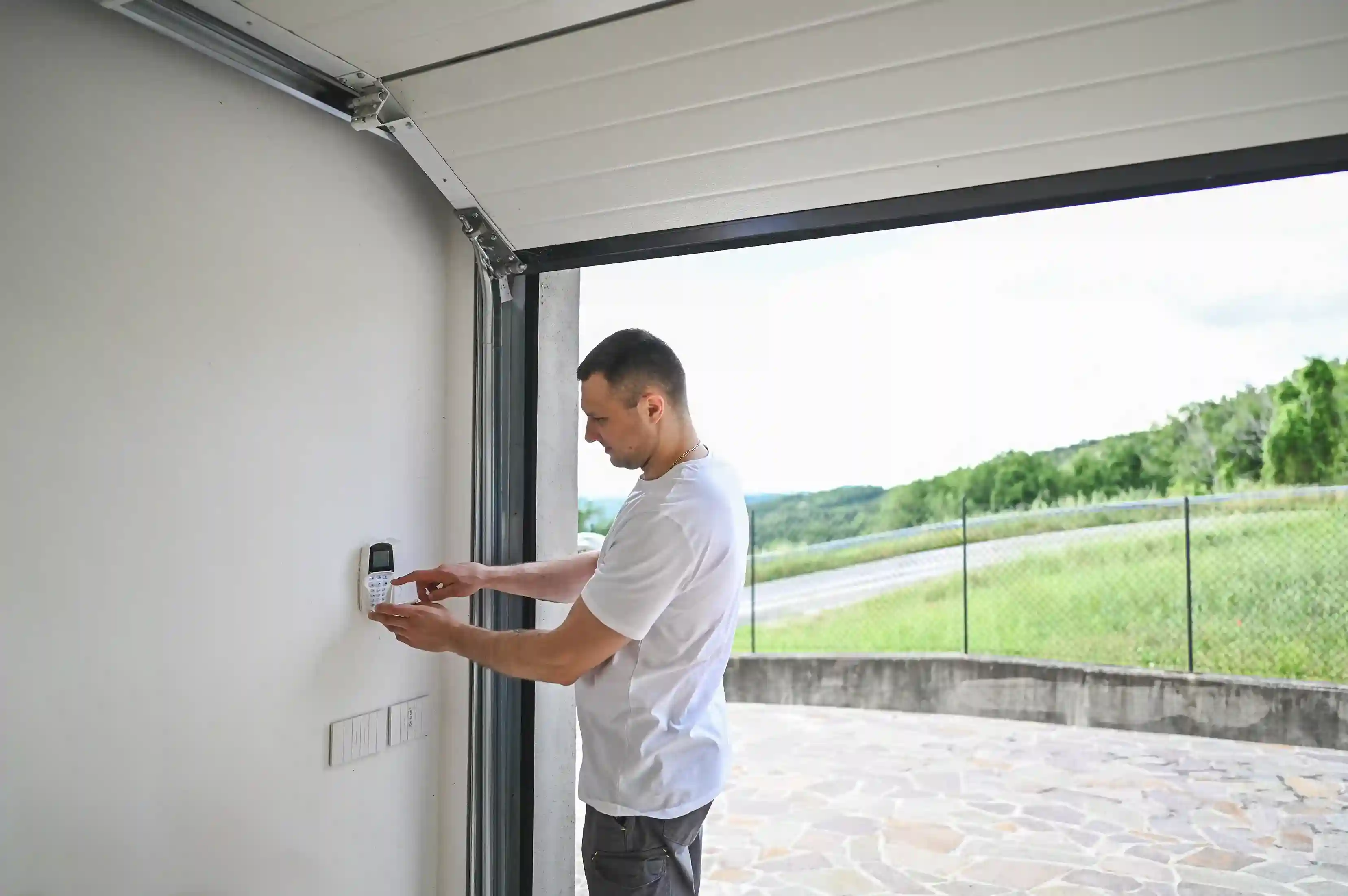 A man in a white t-shirt entering a code on a keypad mounted on the wall inside an open garage, with a view of a grassy landscape outside.