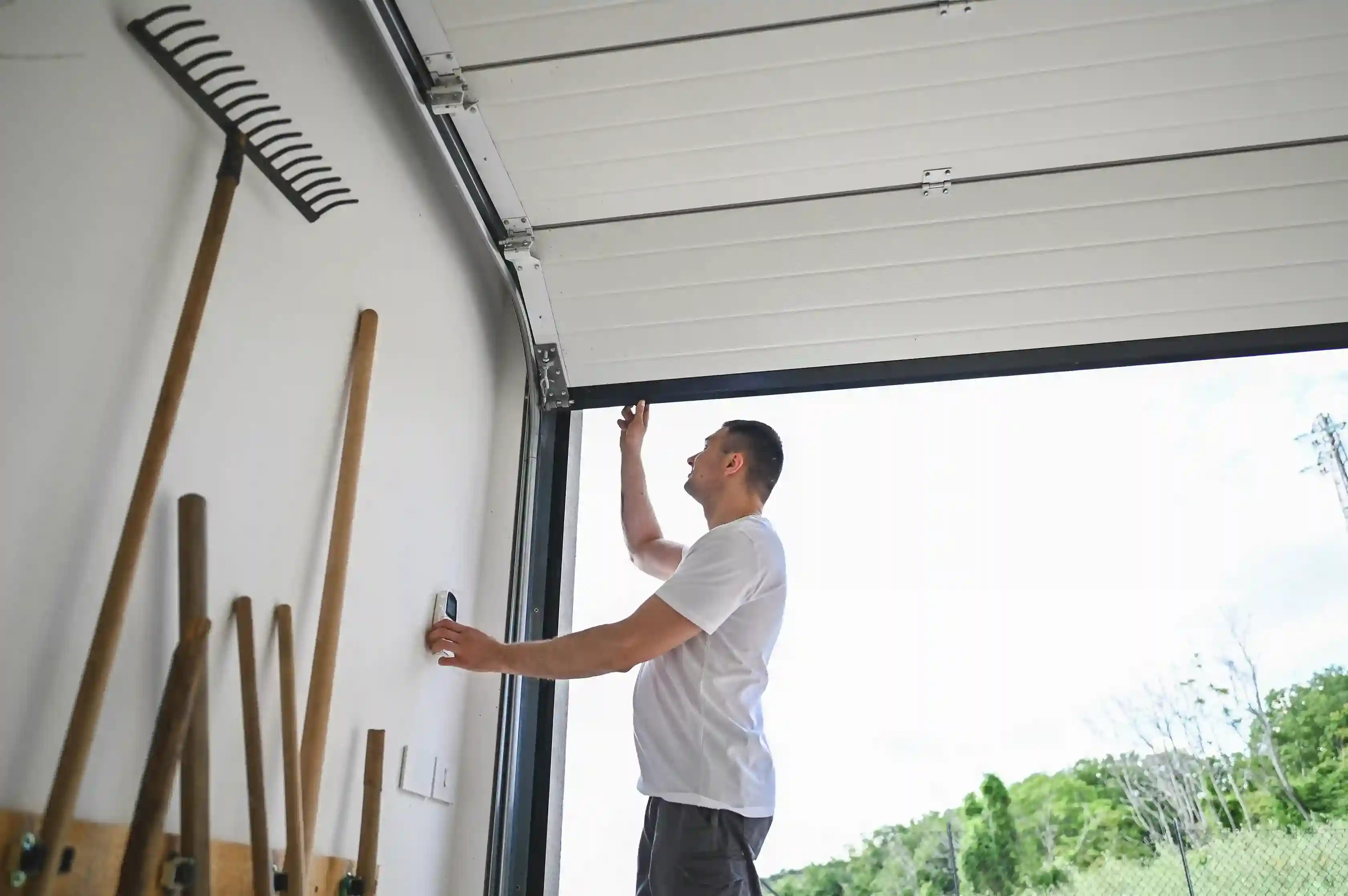 A man in a white t-shirt reaching up to the garage door mechanism from inside an open garage, with rakes hanging on the wall.