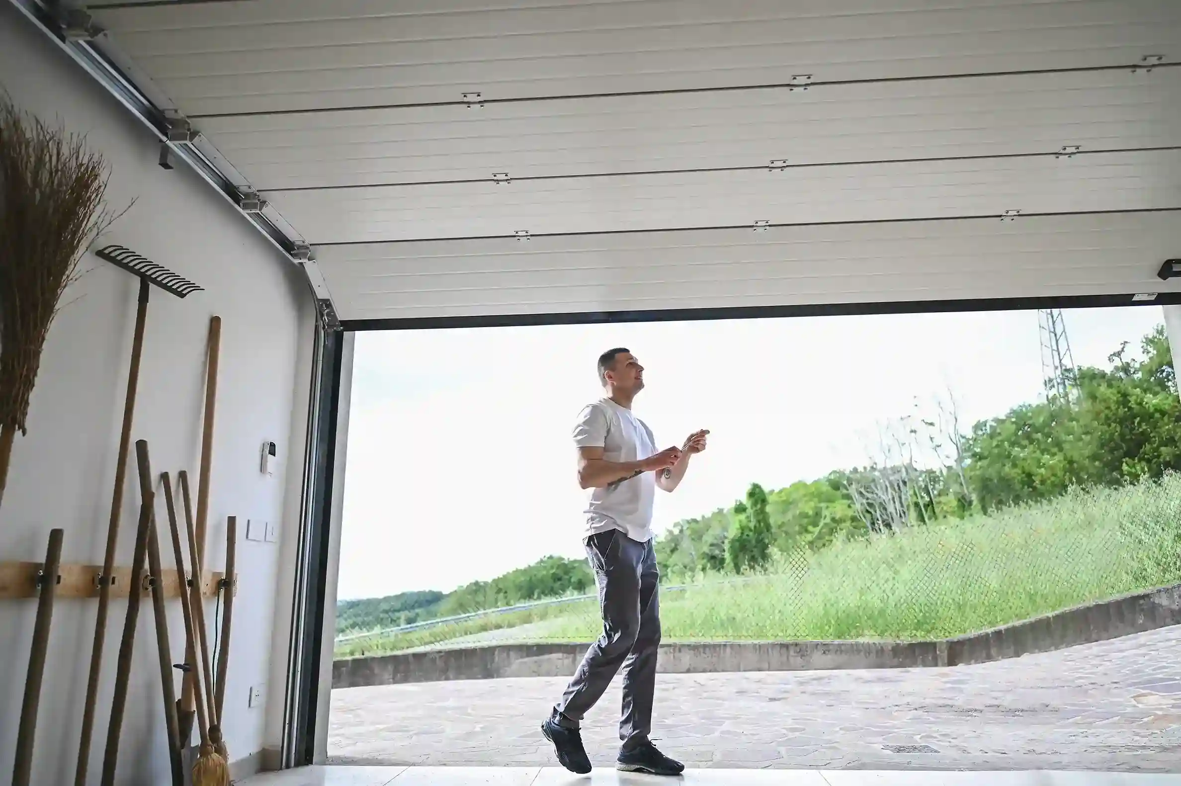 A man in a white t-shirt walking through an open garage, holding a garage door remote, with a view of a grassy hillside outside.