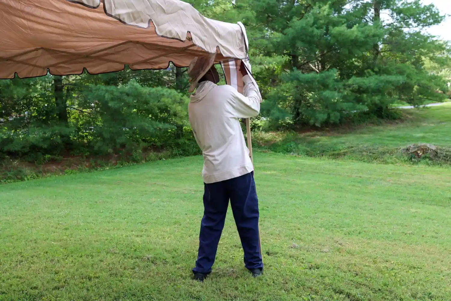 A person in a light-colored long-sleeved shirt and blue pants is standing on a green lawn, holding up a large, light brown fabric awning structure.