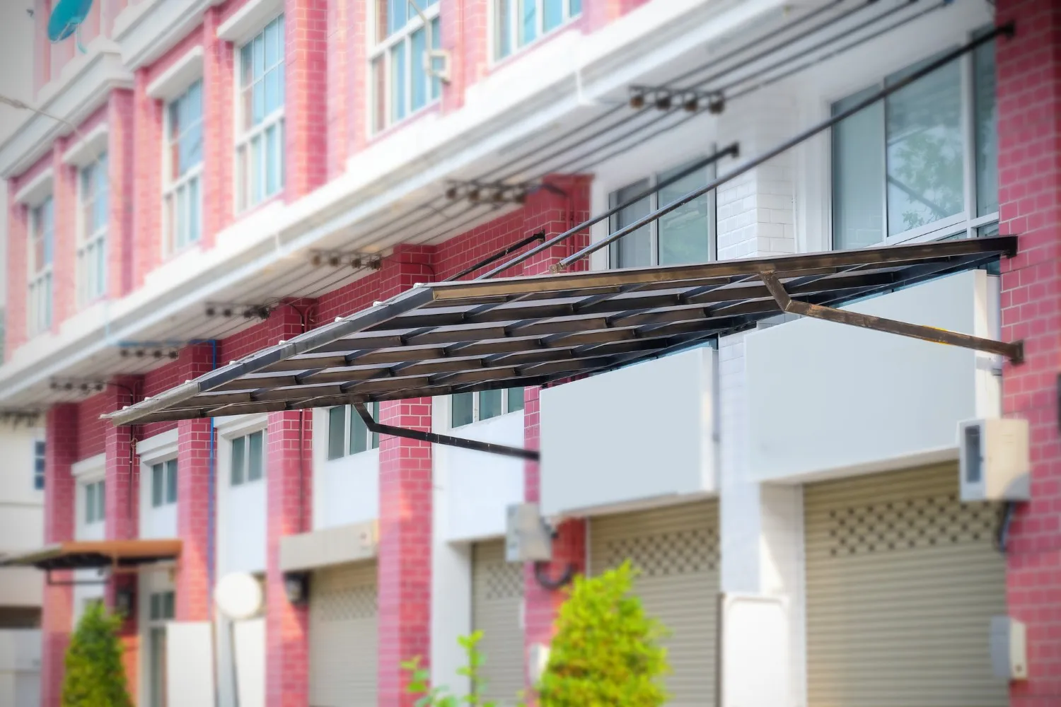 A new dark gray metal awning is being installed on a building with red brick and white sections, featuring multiple windows and roll-up garage doors.