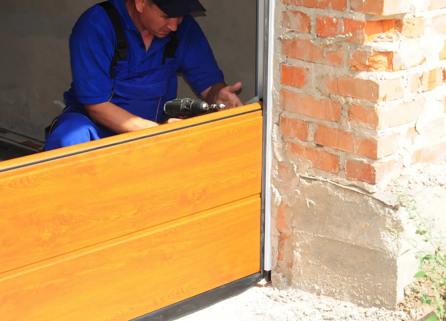 A technician in blue overalls and a cap is using a power drill to install or repair a section of a wooden-paneled garage door, with a brick wall visible on the right.