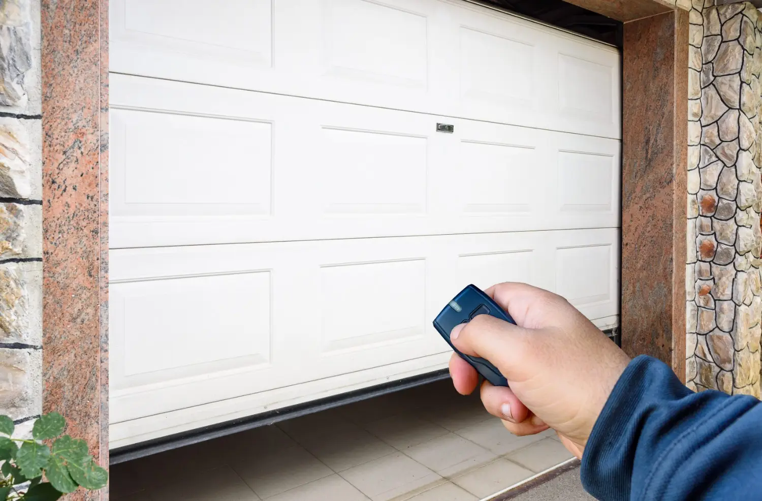 A person's hand is holding a black garage door opener remote, pointing it towards a partially open white paneled garage door framed by stone pillars.