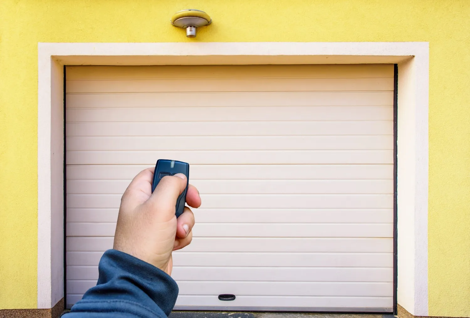 A person's hand is holding a black garage door remote, pointing it towards a closed beige garage door set into a yellow wall with white trim.