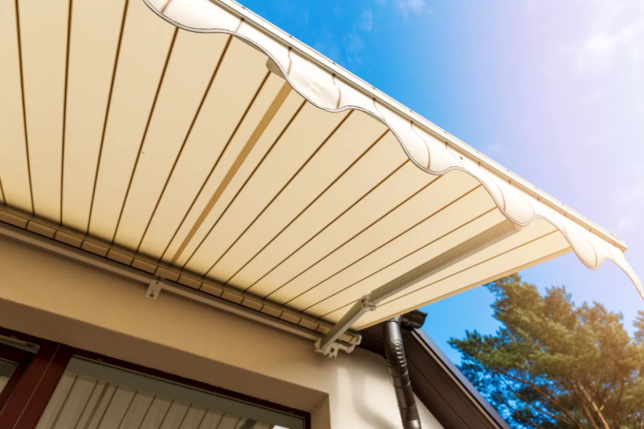 A low-angle shot of a beige-striped awning with a scalloped edge against a bright blue sky, attached to a house with a brown roof.