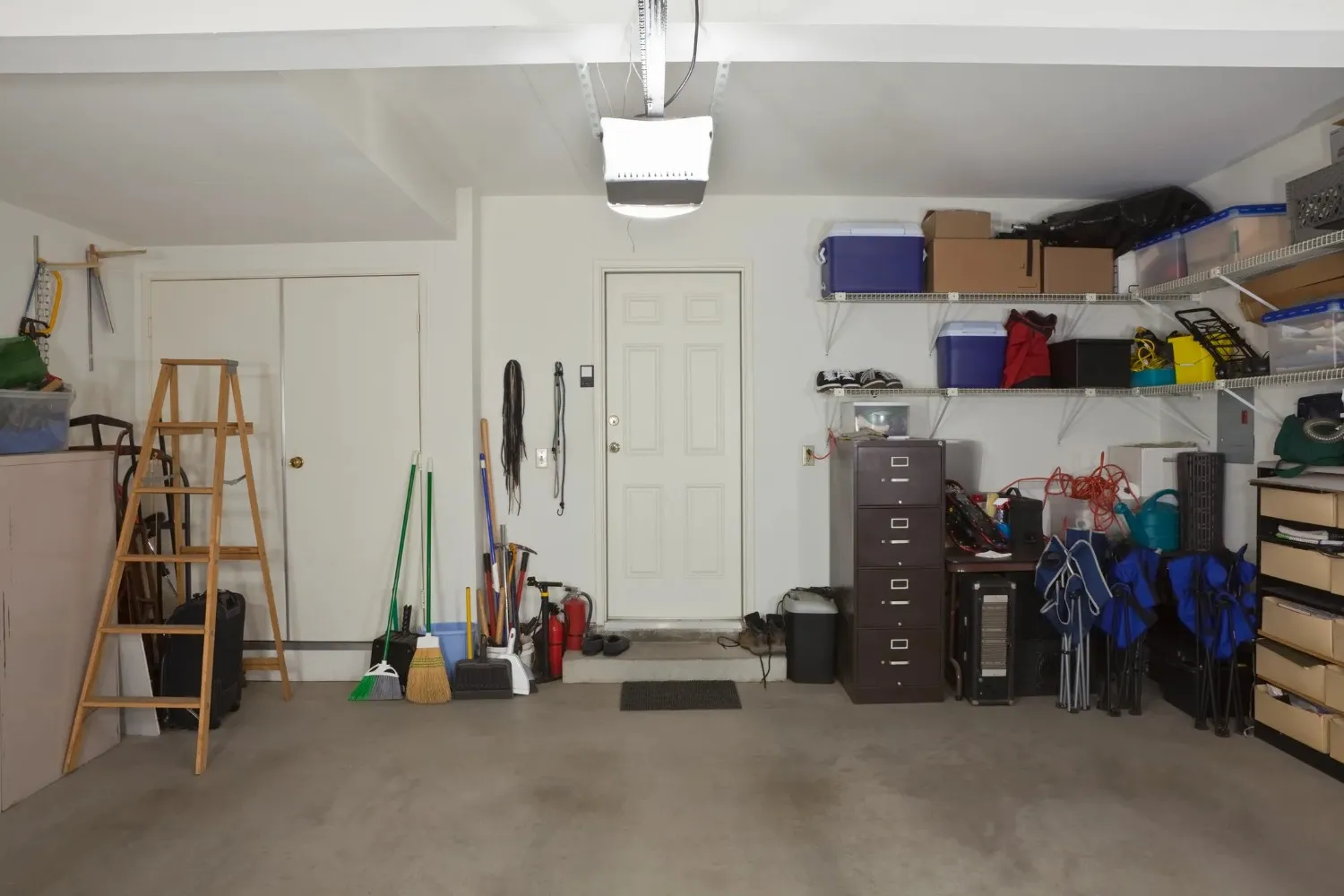 An organized garage interior with two large shelving units on the right, a work ladder, and a closed door, all beneath a ceiling-mounted garage door opener.