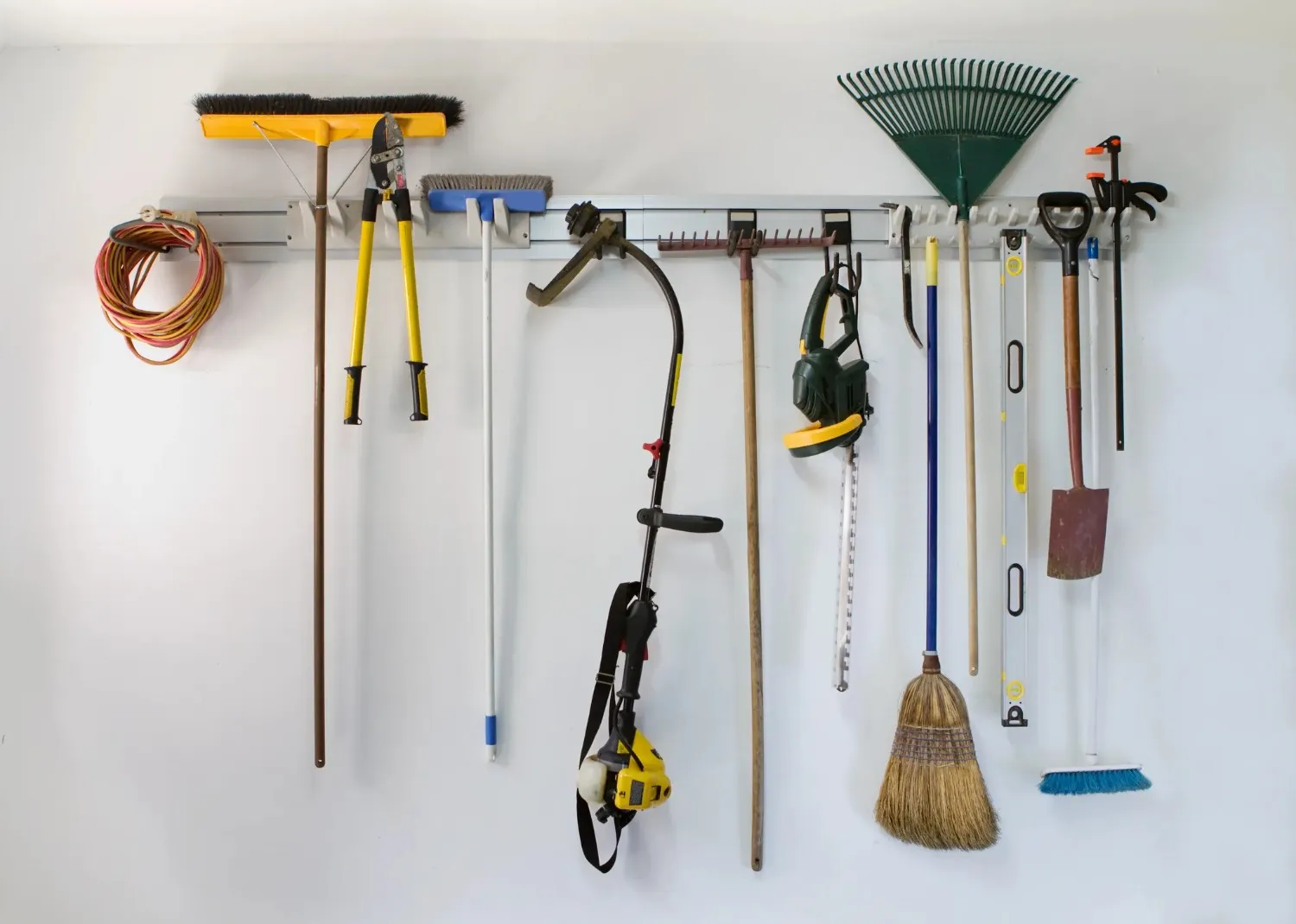 A close-up of a wall-mounted garage storage system holding a variety of long-handled tools, including brooms, rakes, and a weed trimmer.