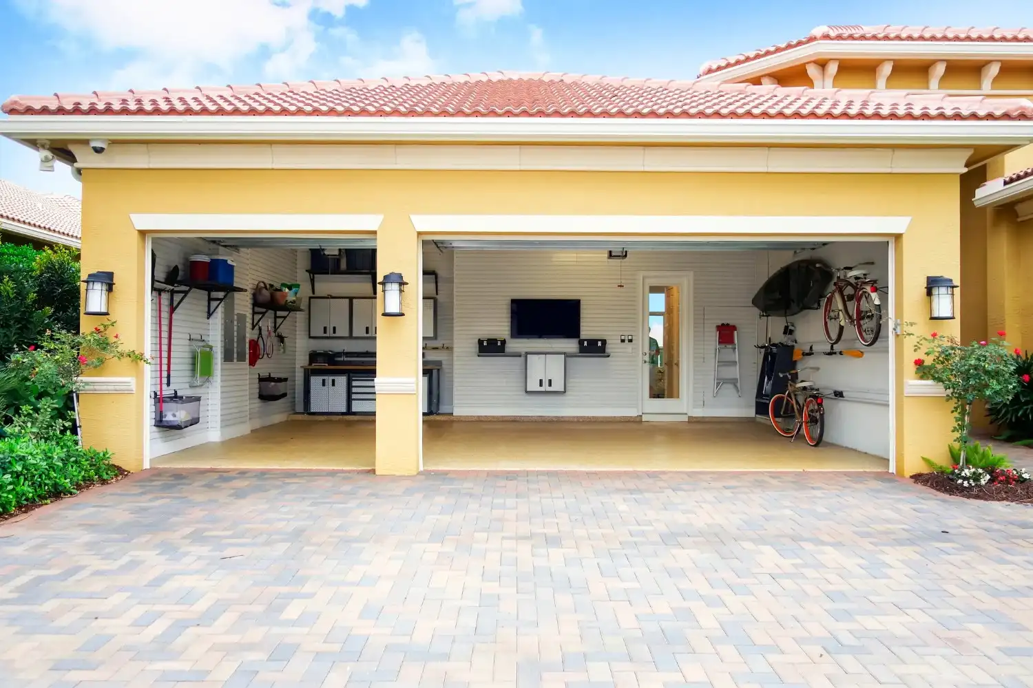 An exterior view of a two-car garage with a paver driveway, showing an organized interior with wall-mounted bikes and a television.
