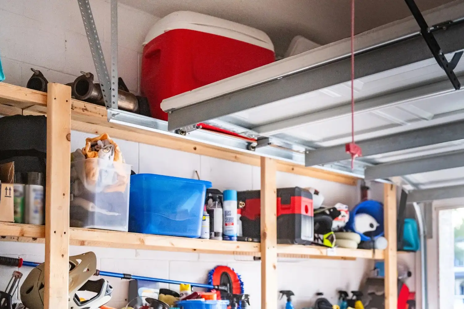 A close-up view of wooden overhead garage shelving, showing a red and white cooler and various storage boxes and items, with the garage door mechanism visible above.