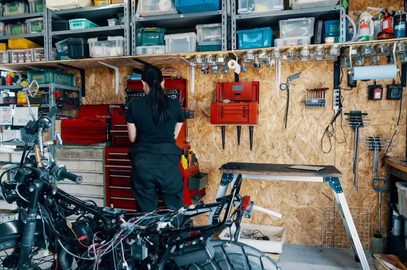 A person in black clothing works on a motorcycle in a cluttered garage with a pegboard wall, metal cabinets, and open shelving filled with tools and parts.