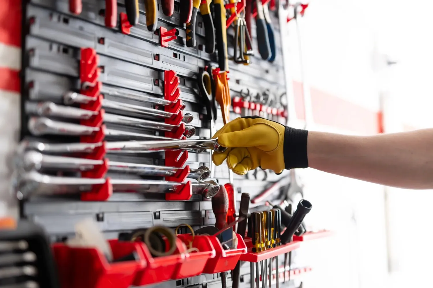 A close-up shot of a hand in a yellow glove reaching for a wrench from a red and black wall-mounted tool organizer.