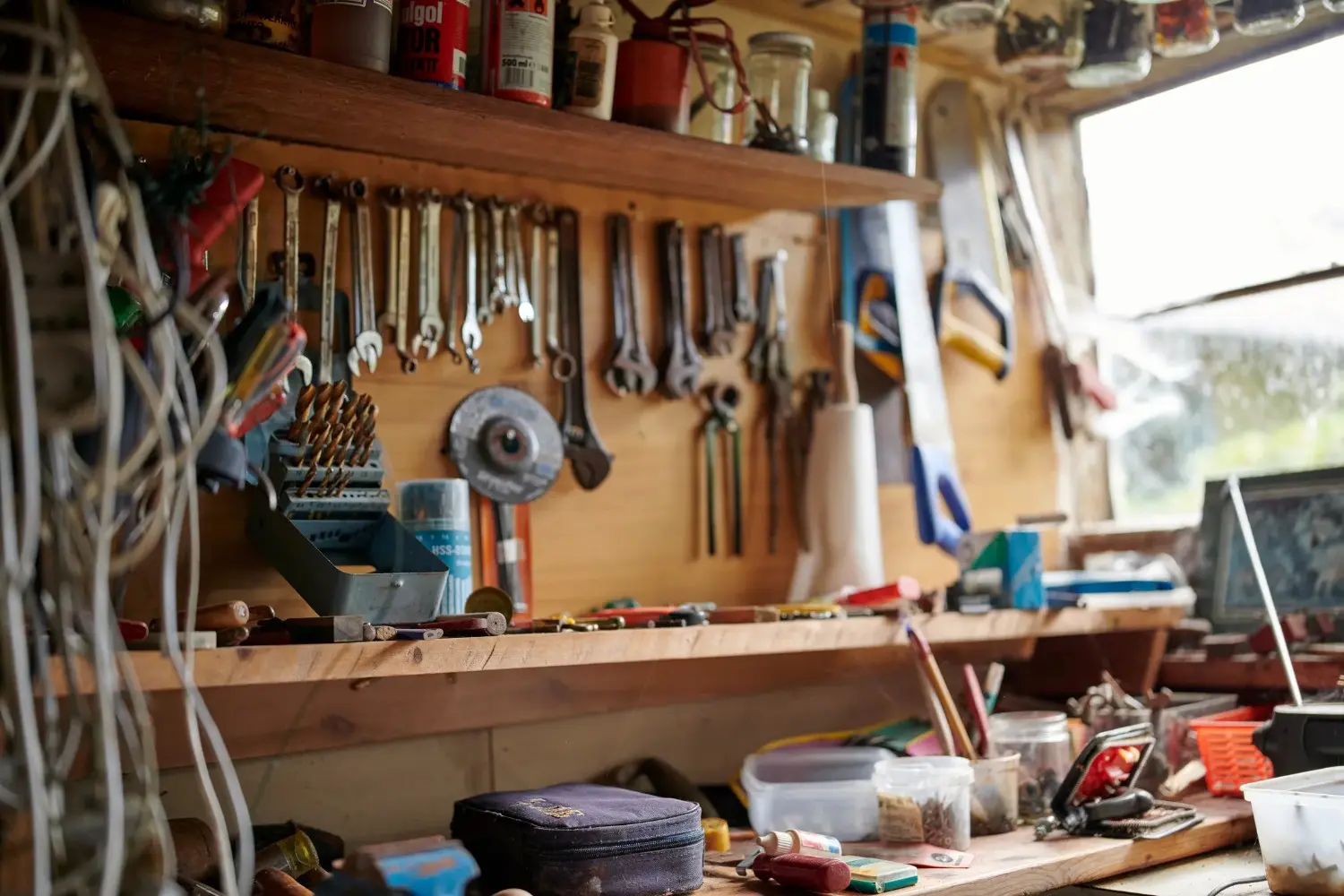 A close-up view of a cluttered wooden workbench in a workshop, with various tools, cans, and a window in the background.