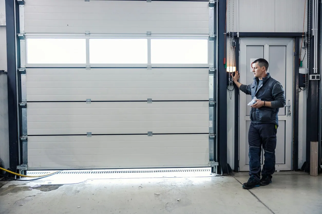 Person in work attire presses control panel button beside large segmented industrial garage door inside facility.