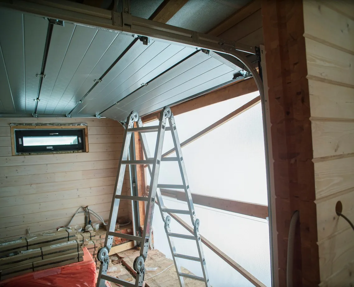 Interior of wooden garage under construction with metal ladder leaning on partially open sectional door.