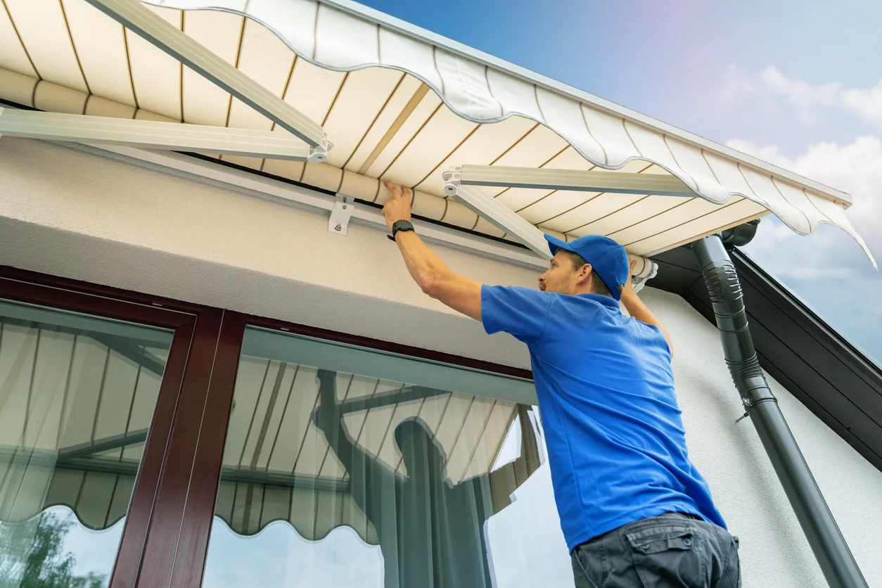 A worker in a blue shirt and cap adjusts a beige retractable awning above a window