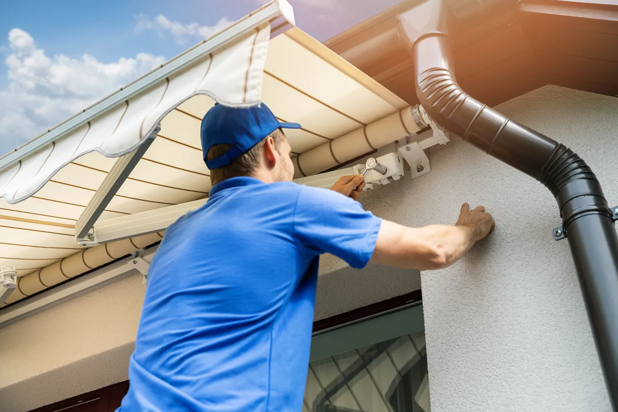 A technician in a blue cap installs a white awning mechanism on a home's exterior wall
