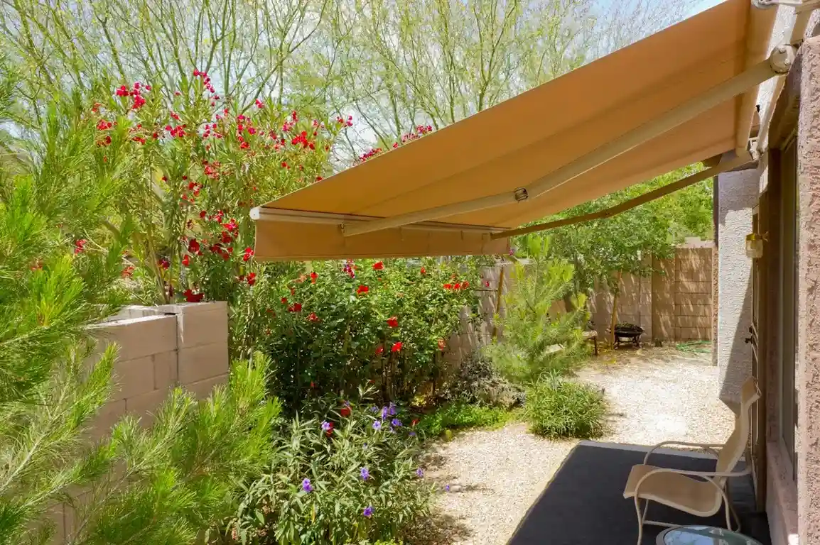 A beige retractable awning provides shade over a patio area in a sunny, landscaped backyard with a stone wall and various flowering plants.