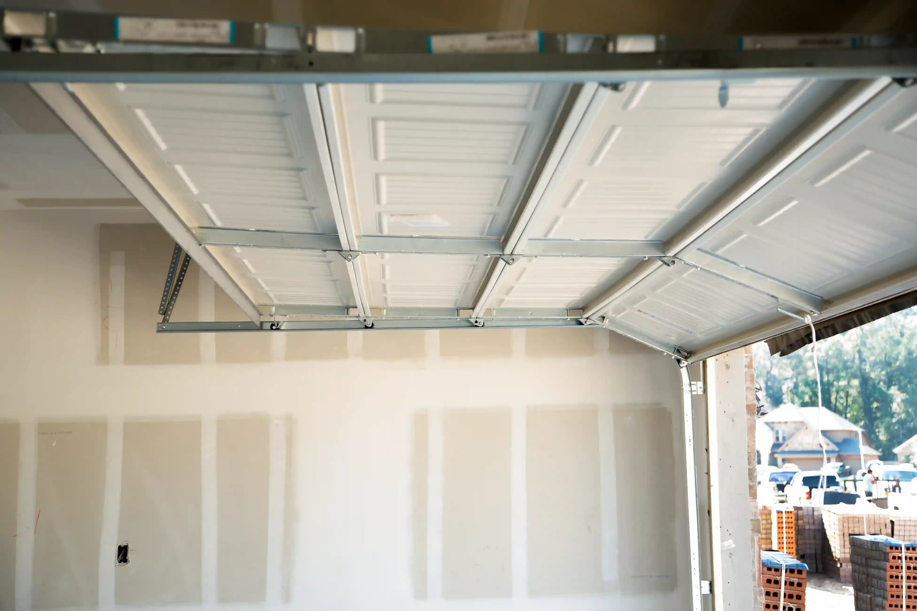 An inside view of a white paneled garage door opening upwards, revealing an unfinished garage interior and a construction site outside.