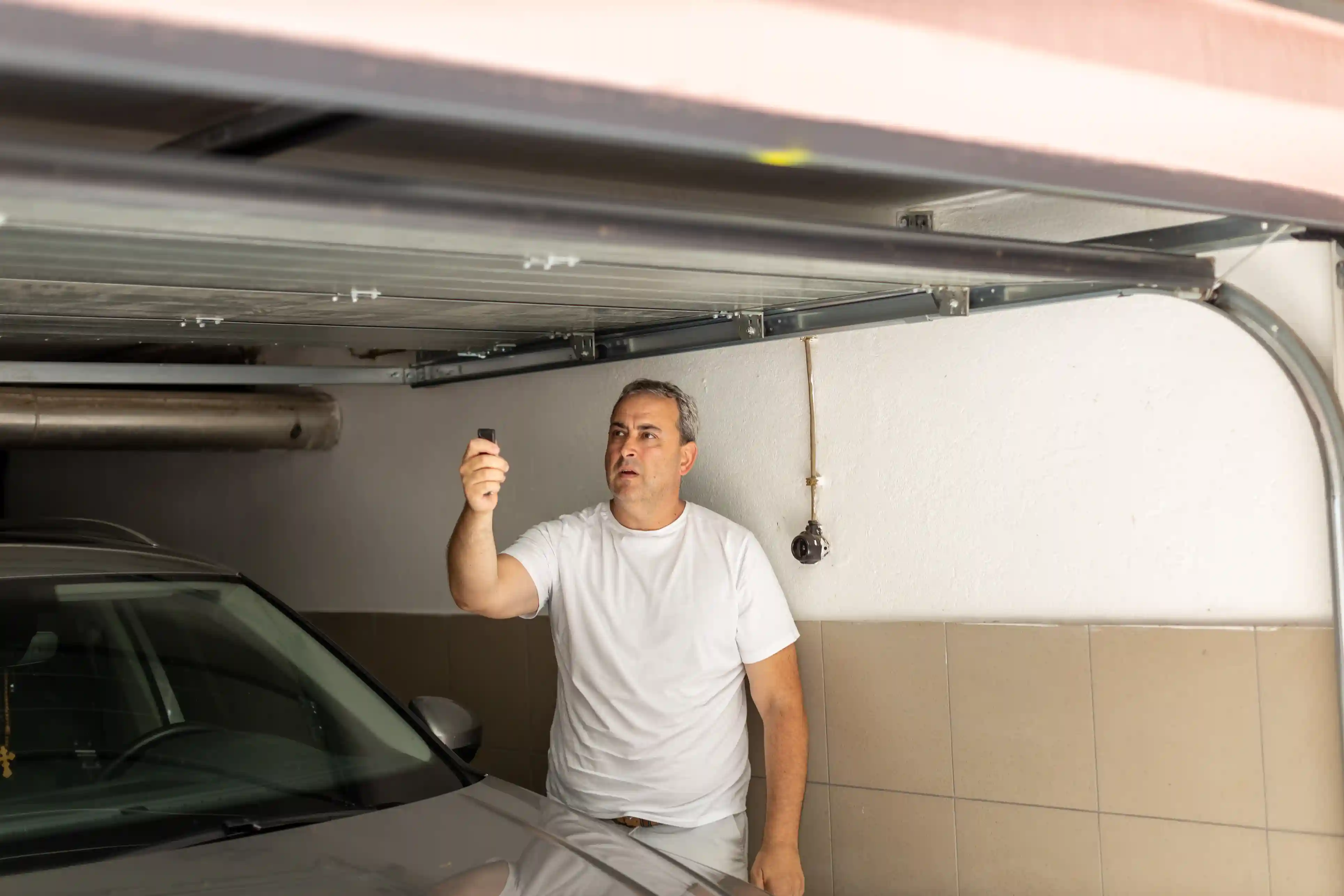 A man in a white t-shirt operating a garage door opener remote from inside his garage, with a car visible in the foreground.