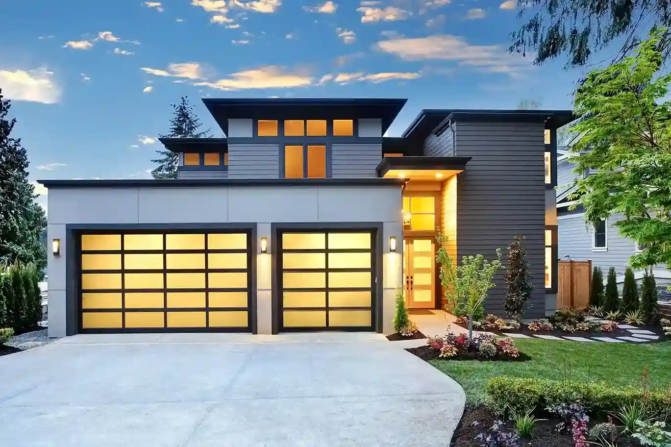 A modern house with a gray and white exterior and two garage doors featuring large frosted glass panels with black frames, illuminated from within.
