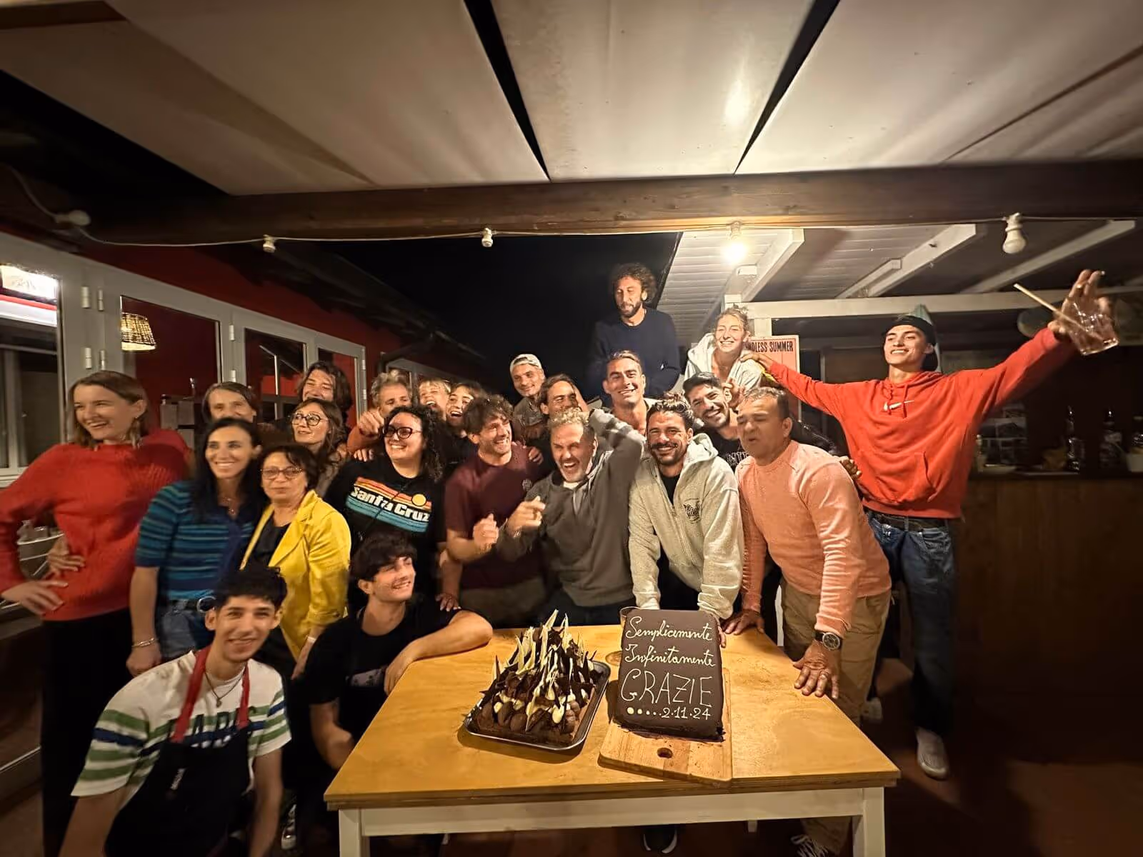 Group of smiling people gathered around a table with two chocolate cakes, one with a thank you message in Italian dated 2.11.24.