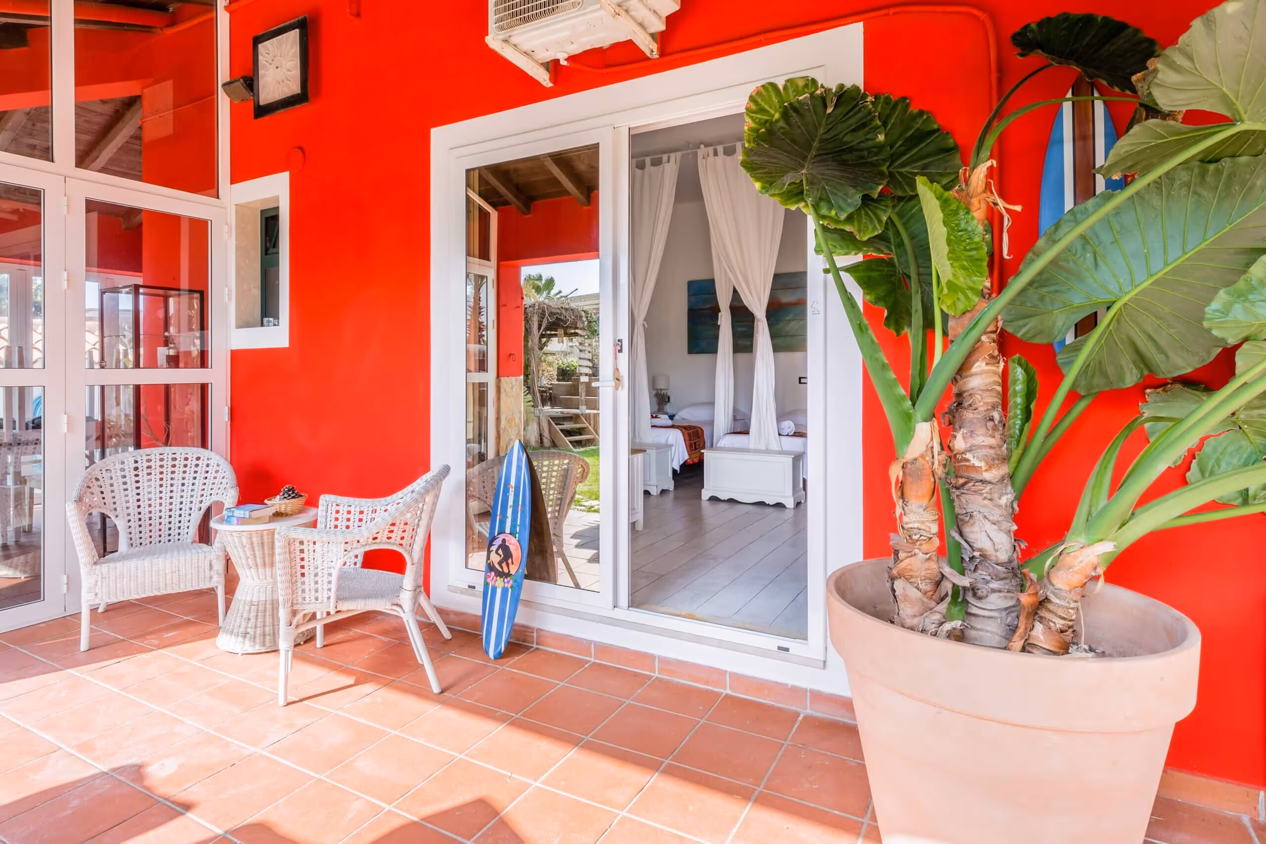 Outdoor patio with two white wicker chairs and table, large potted plant, blue surfboard, and sliding glass doors leading to a bedroom with white curtains and beds.
