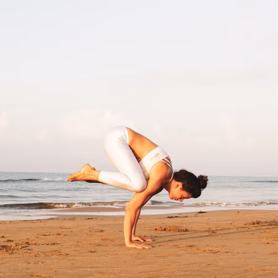 Woman in white activewear performing a yoga crow pose on the sandy beach near the ocean.