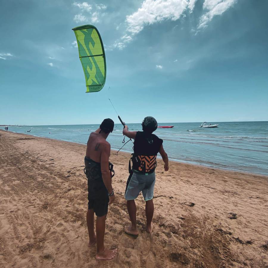 Kitesurf instructor teaching a student on the beach at Floripa Surf and Yoga Retreat in Sicily, with a green kite flying under a cloudy sky