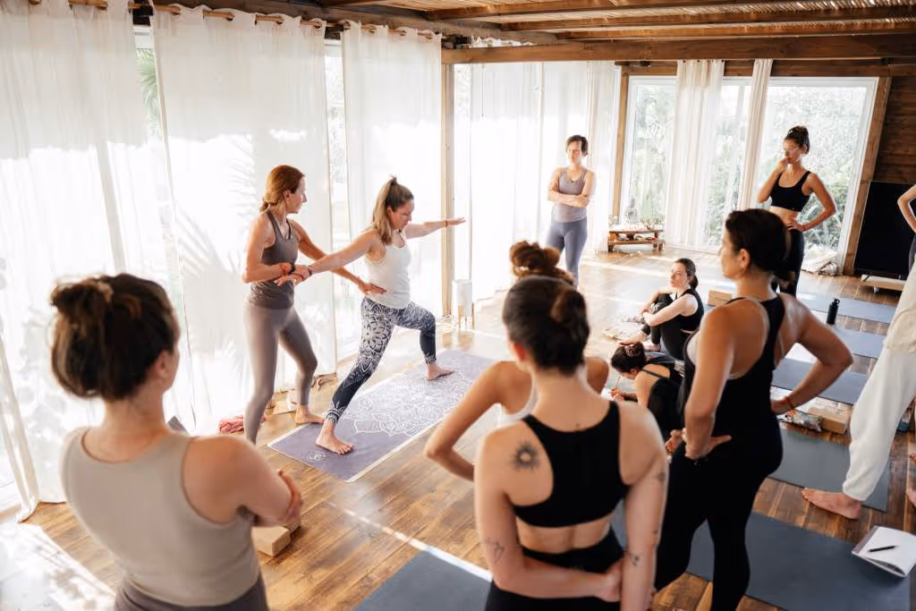 A yoga instructor guides a woman in warrior pose while a group of people watch inside a sunlit studio with wooden floors and white curtains.