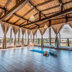 Spacious yoga studio with wooden floor, large windows draped with beige curtains, and yoga mat with blocks and a strap.