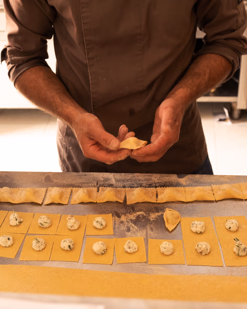 Person shaping homemade stuffed pasta with dough squares and filling on a floured surface.
