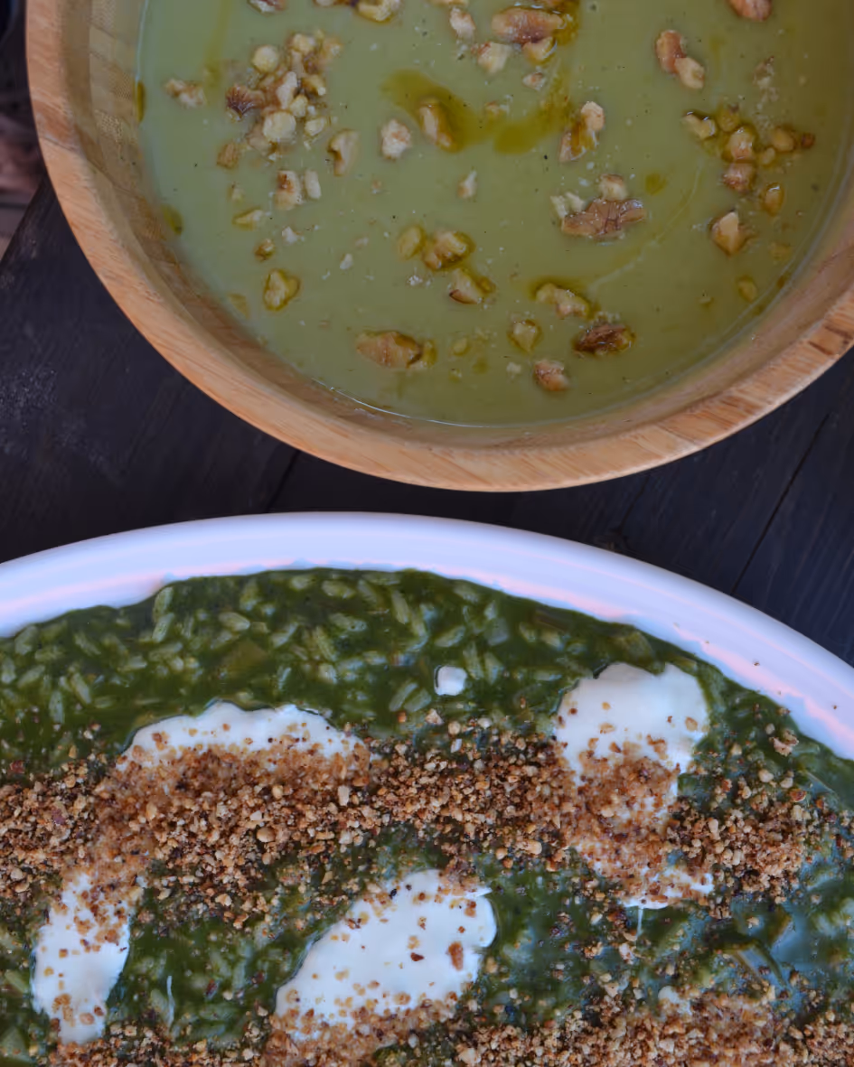 Top view of a wooden bowl with green soup garnished with walnut pieces and a white dish with green creamy spinach rice topped with white sauce and crushed nuts.