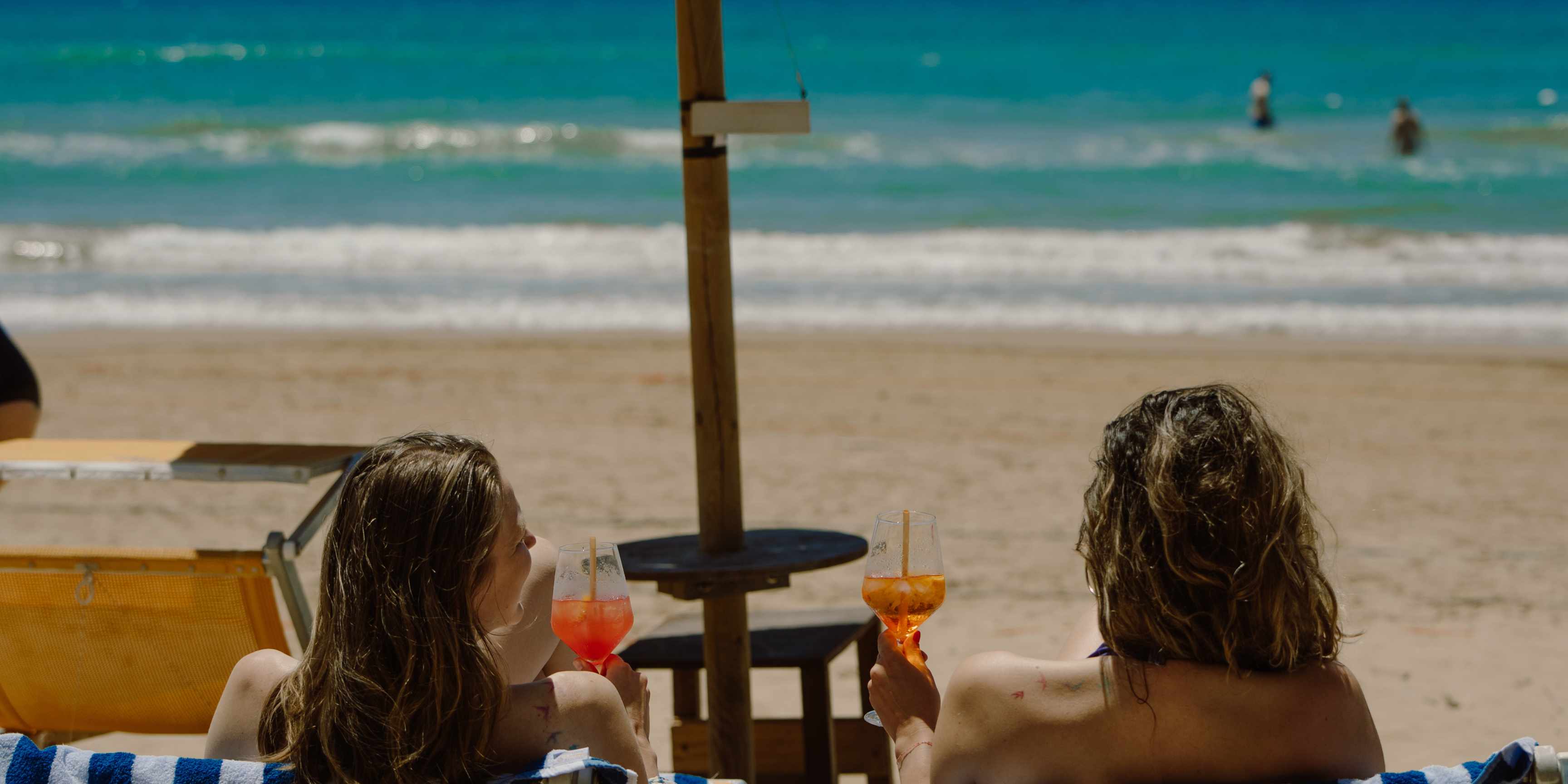 Two guests enjoying drinks together on the beach at Floripa Surf and Yoga Retreat in Sicily, zuma beach, embodying the spirit of connection and community