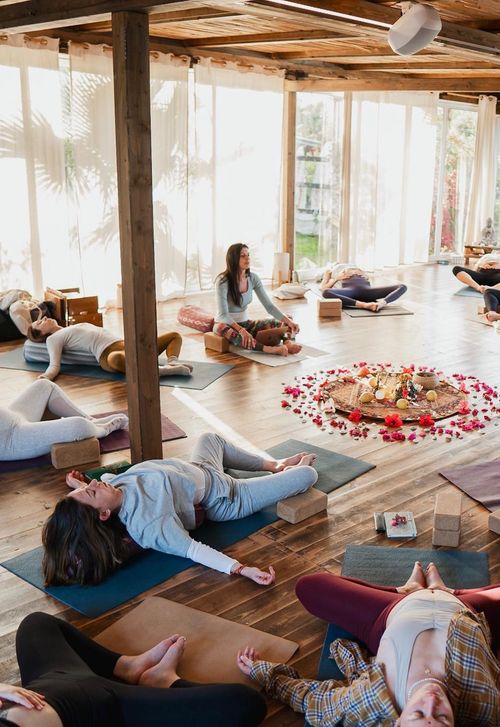 Group of people practicing yoga in a bright room with wooden floors and large windows, some lying down and one sitting cross-legged near a floral mandala arrangement.