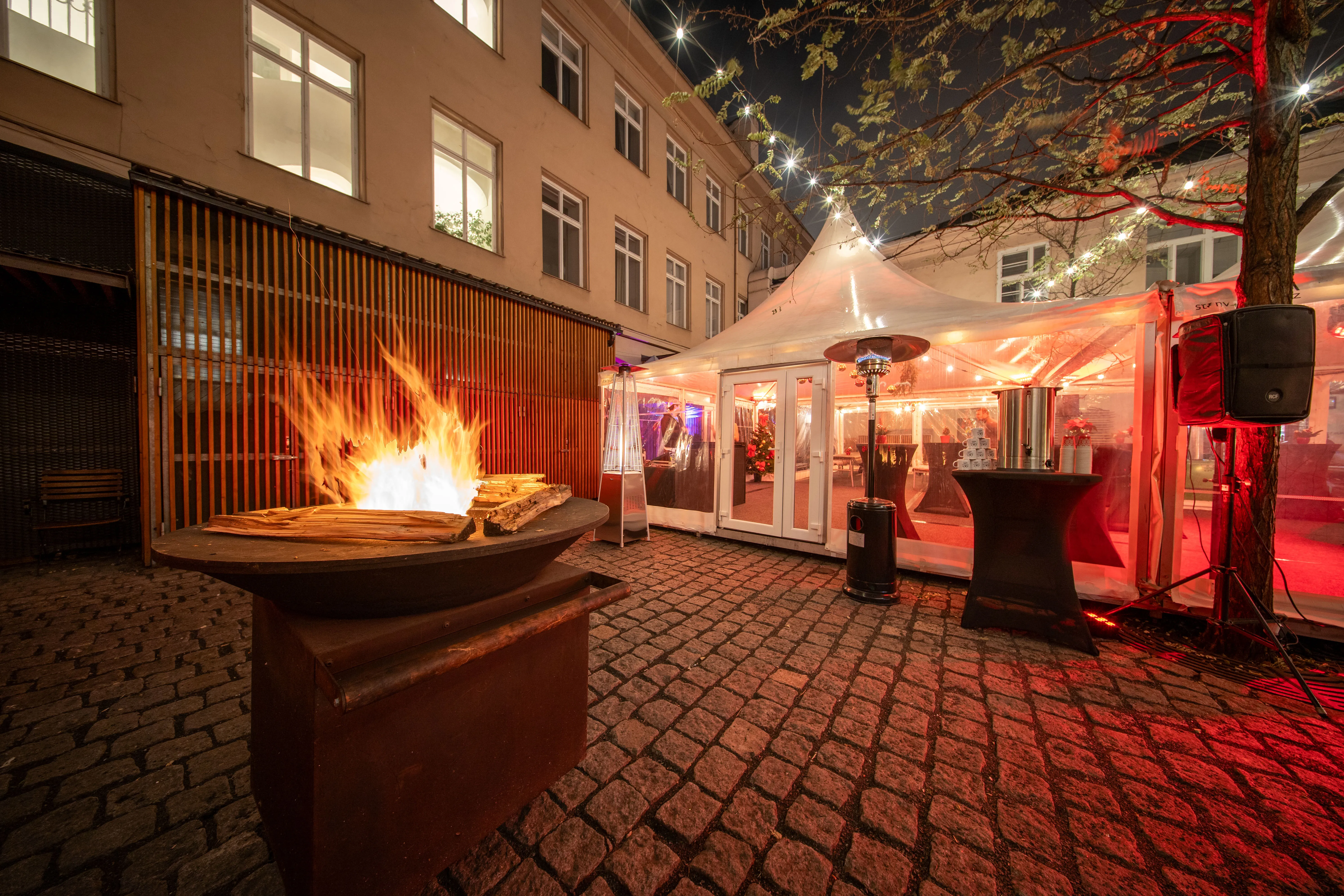 Venkovní prostor s ohništěm, osvětleným stromem a proskleným stanem při noční akci.