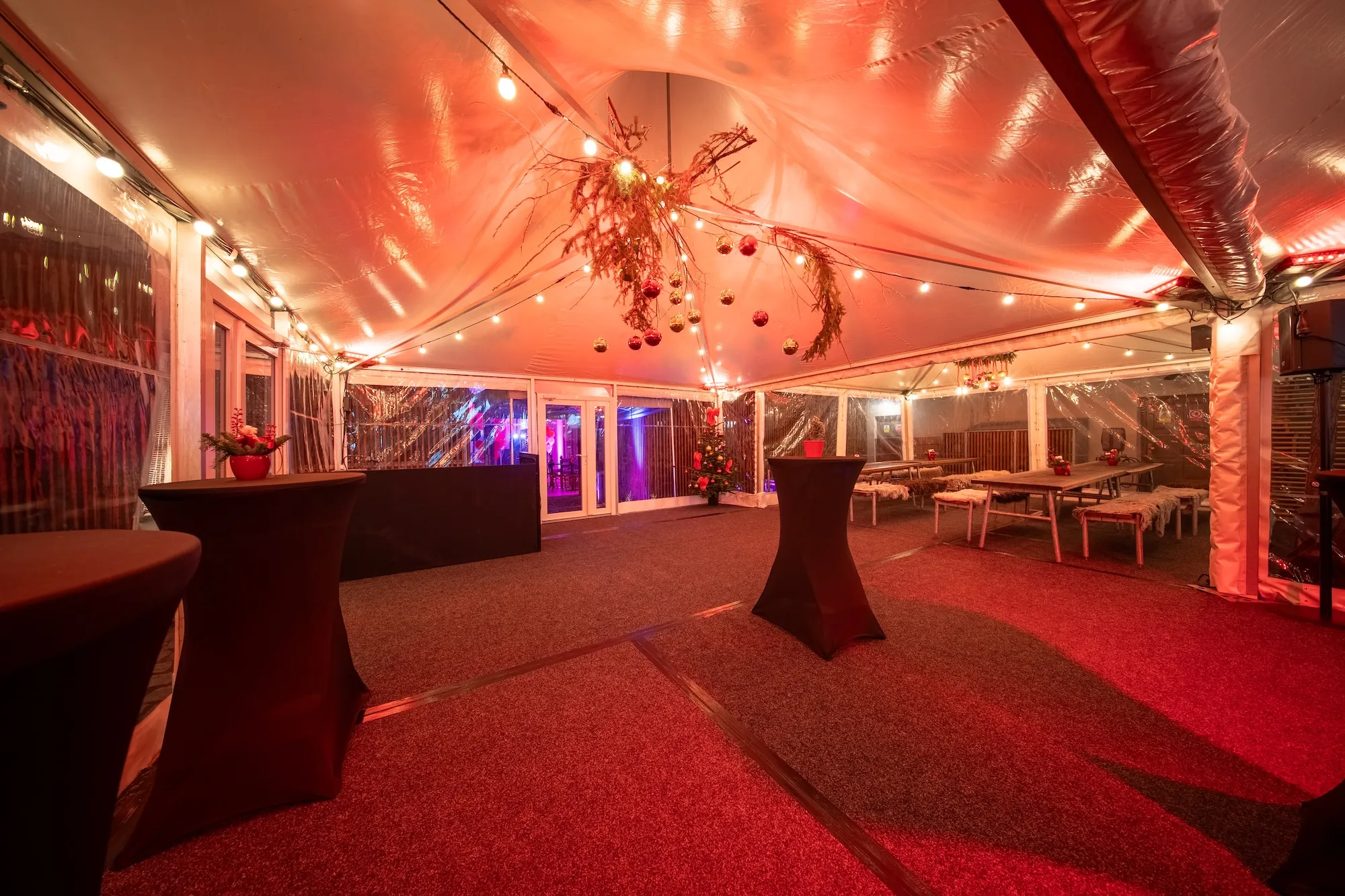 Osvětlený interiér velkého stanu s vánoční výzdobou, včetně ozdob na stropě a stolů pokrytých černými potahy.